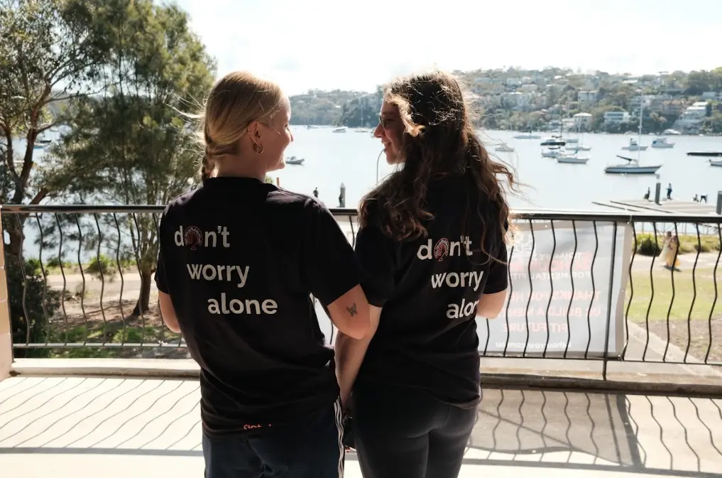 Two women in “don’t worry alone” shirts stand together on a balcony overlooking a sunny harbor, boats, and hillside homes.