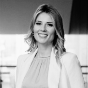 Black-and-white portrait of a smiling woman with shoulder-length hair, wearing a light blazer and blouse, posed against a bright indoor background; warm, professional tone.